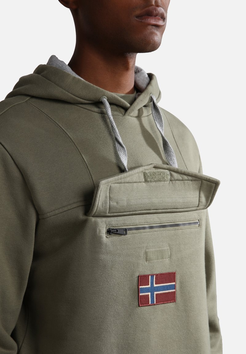 Homme portant un sweat à capuche vert olive avec des cordons gris et une poche avant avec rabat zippé et un écusson du drapeau norvégien.
