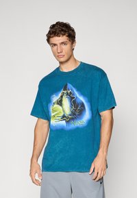 Ung mand iført en blå oversized t-shirt med et grafisk motiv af en skægget troldmand, der holder en lysende basketball og teksten "Nike".