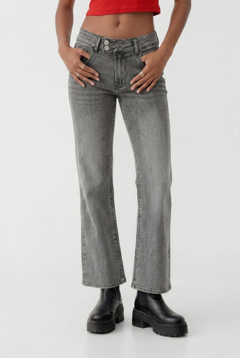 Femme portant un jean gris délavé taille haute avec des jambes évasées, des bottines noires épaisses et un top court rouge, mains dans les poches avant.