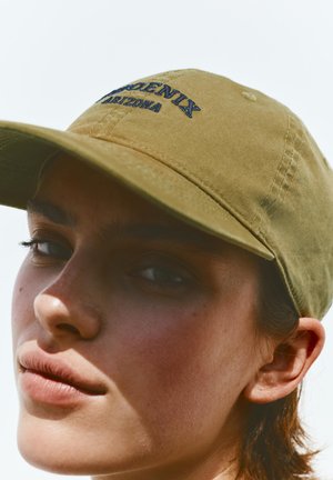 Kobieta nosząca beżową czapkę z daszkiem z niebieskim haftem „Phoenix Arizona”, zwrócona lekko w lewo, z neutralnym wyrazem twarzy i naturalnym oświetleniem.