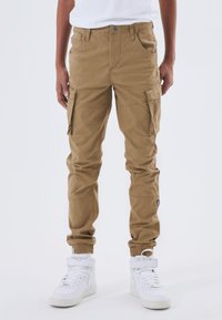 Pantaloni cargo marroni con polsini elasticizzati, dotati di numerose tasche e patta con zip. Indossati con sneakers bianche alte.