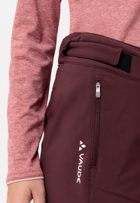 Bordeauxrode broek met een hoge taille, voorzien van een zijzak met rits en een wit logo. Vergezeld van een rood textuur langemouwen top.