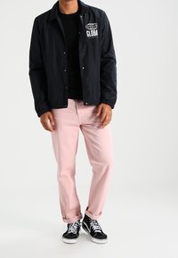 Man in zwarte jas met "GLOBAL" logo, zwart shirt, lichtroze broek en zwarte high-top sneakers staat op een witte achtergrond.