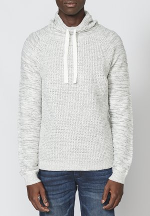 Pull gris clair avec col châle et cordons. Texture tricotée, poignets et ourlet côtelés, présentant une coupe légèrement ample. Associé à un jean foncé.