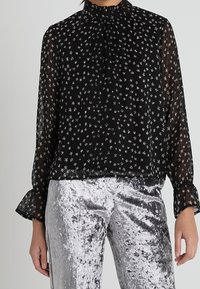 Schwarze, transparente Bluse mit hohem Kragen, die ein kleines Blumenmuster in Weiß aufweist. Lange, ausgestellte Ärmel, getragen mit silbernen Samthosen.