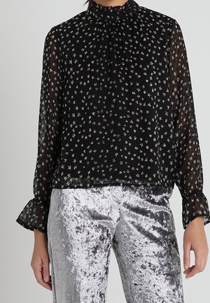 Schwarze, transparente Bluse mit hohem Kragen, die ein kleines Blumenmuster in Weiß aufweist. Lange, ausgestellte Ärmel, getragen mit silbernen Samthosen.
