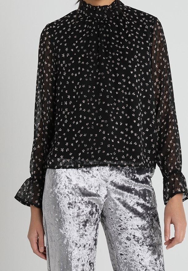 Schwarze, transparente Bluse mit hohem Kragen, die ein kleines Blumenmuster in Weiß aufweist. Lange, ausgestellte Ärmel, getragen mit silbernen Samthosen.