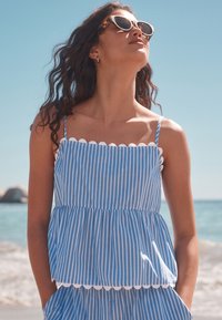 Blå og hvide stribede tanktop med bølgetudskæring og peplum-design. Letvægtsstof, tynde skulderstropper og afslappet pasform.