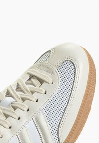 Baltas ādas sporta apavs ar acu paneļiem, apaļu purngalu un plakaniem auklīšu caurumiem. Iekļauj gumijas zoli un kontrastējošas šūšanas detaļas.