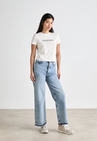 Biała bawełniana koszulka z napisem "Calvin Klein Jeans", połączona z jasnoniebieskimi szerokimi spodniami dżinsowymi i szarymi trampkami. Prosty, casualowy zestaw.