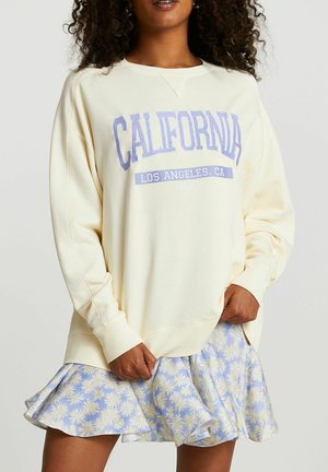 Femme portant un sweat crème avec l'inscription "CALIFORNIA LOS ANGELES, CA USA" en violet, associée à une jupe à volants à fleurs bleues et blanches.