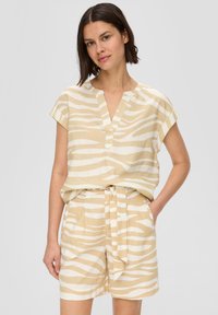 Junge Frau, die ein beige-weißes, gestreiftes Kurzarmoberteil trägt, das in passende hochtaillierte Shorts mit gebundenem Gürtel gesteckt ist, steht vor einem grauen Hintergrund.