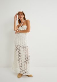 Weißes Häkel-Maxi-Kleid mit Halterneck, zentralem Knotendetail und einem offenen Muster mit Blattformen. Akzentuiert mit beigen Schuhen.