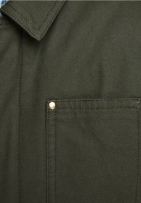 Gros plan d'une poche de chemise en tissu vert olive avec un rivet en laiton et des coutures apparentes près du col.