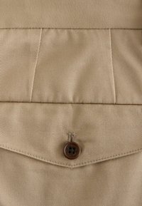 Gros plan sur un tissu beige avec une poche à rabat fermée par un unique bouton brun foncé à quatre trous.