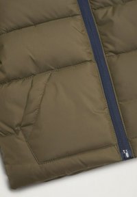 Oliwkowa pikowana kurtka z zaznaczonym skośnym bocznym kieszeniem i niebieskim zamkiem na tle białym.