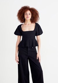 Top peplum nero con motivo zigzag testurizzato, scollatura quadrata e maniche a sbuffo corte, abbinato a pantaloni neri a vita alta e dalla gamba ampia.