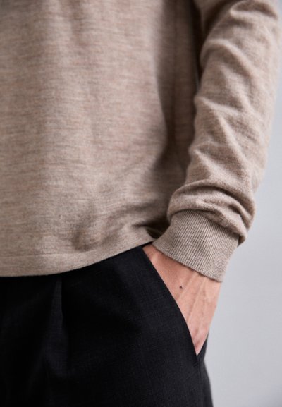 Maglione lavorato a maglia beige con polsini a coste, abbinato a pantaloni scuri, che presenta una mano in tasca. Texture liscia, design semplice.