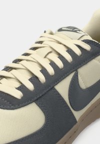 Sneaker beige con accenti in suede grigio, lacci beige e suola in gomma. Presenta un logo Nike prominente sulla linguetta e sul lato.