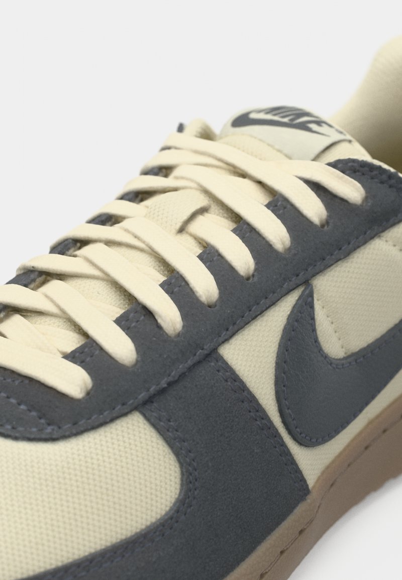 Beige sneaker met grijze suède accenten, beige veters en een rubberen zool. Beschikt over een prominent Nike-logo op de tong en zijkant.