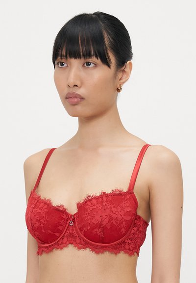Red lace balconette bra featuring floral patterns, satin straps, scalloped edges, and a small decorative accent at the center.