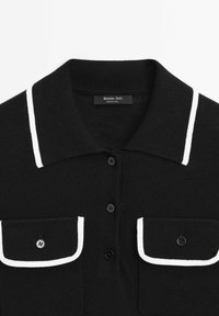 Chemise noire à col avec bordure blanche sur le col et les rabats des poches, trois boutons noirs sur le devant, et deux poches poitrine à boutons.