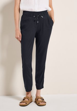 Pantalons légers de couleur marine avec une taille élastique, un cordon de serrage et des poches latérales. Design de jambe fuselée. Portés avec des sandales brunes à enfiler.