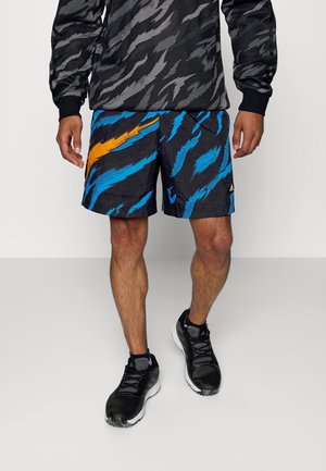 Mand iført sorte sportssko, mønstrede sorte og blå shorts med orange accent, og en sort og grå langærmet top, stående på en hvid gulv.