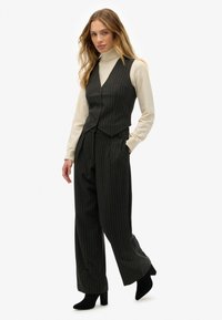 Chaleco de rayas oscuro y pantalones de pierna ancha a juego, hechos de un tejido texturizado. Combinado con un suéter de cuello alto beige claro y botas negras de tacón.