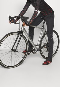 Sølv Triban racercykel med sorte detaljer. Rytter iført sort trøje med røde accenter og cykelsko. Handsker og styr er placeret fremad.