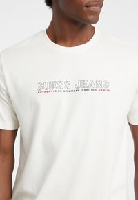 Balta medvilninė marškinėlių su trumpomis rankovėmis ir apvaliu iškirpimu. Pristatomi su spausdintu logotipu "GUESS JEANS" ir tekstu pilka ir raudona spalvomis, einančiu per krūtinę.