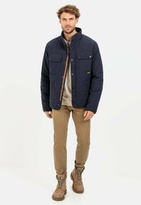 Marineblauer gesteppter Mantel mit zwei Brusttaschen, einem Reißverschluss, der von einer Druckknopfblende verdeckt wird, und einem hellbraunen Kragen. Getragen mit khakifarbenen Hosen und hellbraunen Stiefeln.