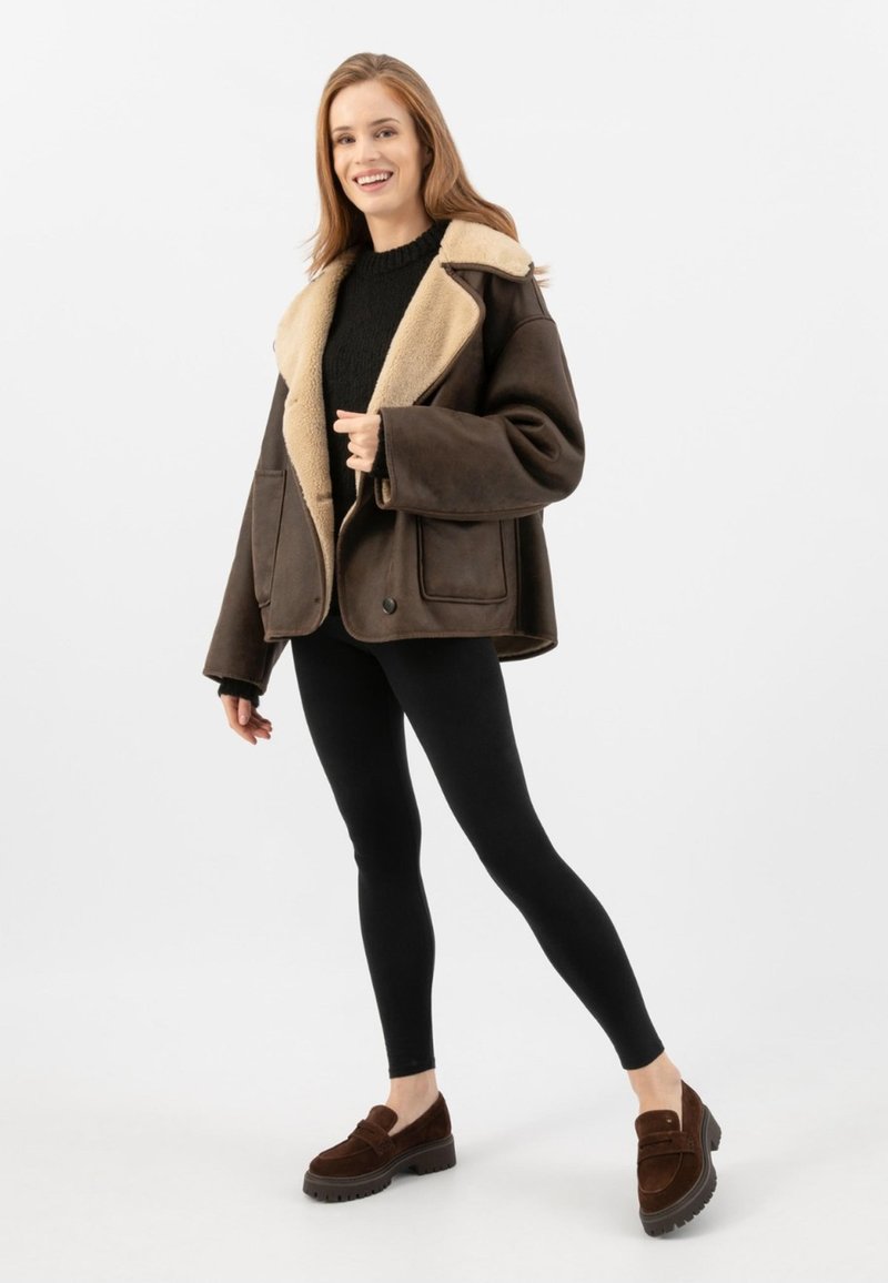 Donna sorridente, vestita con un giubbotto in shearling marrone scuro con rifiniture beige, maglione nero, leggins neri e mocassini marroni, in piedi su uno sfondo bianco.