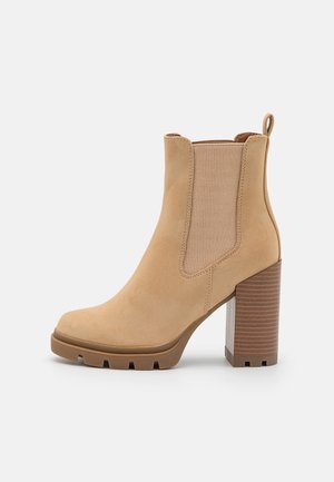 Bottine beige en cuir suédé lisse, avec des panneaux latéraux élastiques et un talon carré affichant une surface brune texturée. Semelle en caoutchouc.