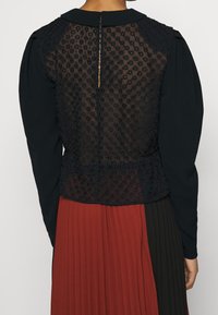 Blusa nera con un retro trasparente a trama floreale. Maniche lunghe, scollatura tonda e una piccola apertura sul retro con chiusura.
