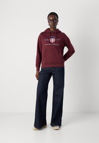 Felpa con cappuccio bordeaux con tasca frontale e design del logo; abbinata a un paio di jeans a gamba larga blu scuro e sneakers beige.