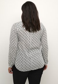 Femme aux cheveux bruns portant un blouse à manches longues noire et blanche et un pantalon noir, se tenant de dos contre un fond uni.