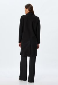Abrigo de lana negro con diseño ajustado, mangas largas, abertura en la parte trasera y corte recto, combinado con pantalones negros de pierna ancha.