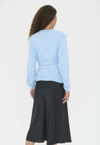 Sieviete ar cirtainiem matiem, ģērbusies vasaras zils džemperī ar jostu vidukļa daļā un melnās vidējās garuma svārkos, stāv uz vienkārša balta fona.