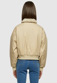 Giacca bomber beige con colletto peloso, maniche oversize e orlo elasticizzato. Tessuto liscio con una finitura opaca.