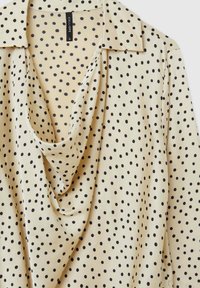 Blouse beige avec un décolleté drapé, des pois noirs et un col classique. Fabriquée en tissu lisse avec une coupe décontractée et fluide.