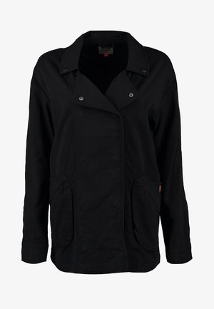 Sort, knap-up jakke med to forlommer, båret over en lys grå skjorte og slidte blå jeans, afsluttet med sorte flade sko.