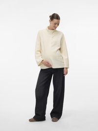 Εγκυμονούσα γυναίκα σε κρεμ πουλόβερ και φαρδιά μαύρα παντελόνια που κρατάει τρυφερά την κοιλιά της, χαμογελώντας και κοιτάζοντας κάτω, όρθια σε λευκό φόντο.