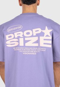Lilla bomulls T-skjorte med store hvite grafikker på baksiden, inkludert ordene "DROP SIZE" og en stjernedetalj, med ekstra tekst under.