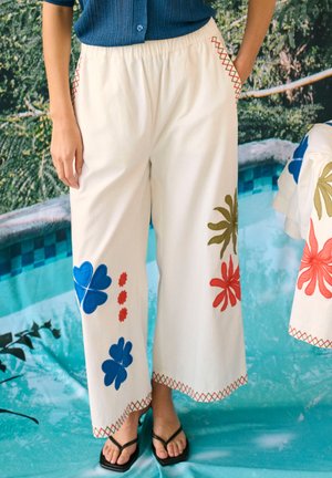 Persona con pantalones anchos blancos con estampados florales azules, rojos y verdes, de pie junto a una prenda a juego sobre un fondo turquesa de piscina.