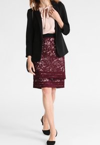 Svart kavaj över en ljusrosa blus, i kombination med en lager-på-lager bordeauxfärgad spetskjol. Svarta klackar fulländar outfiten.