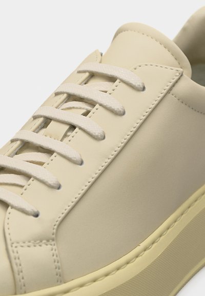 Κοντινό πλάνο ενός μπεζ αθλητικού παπουτσιού με κορδόνια στο ίδιο χρώμα, λεπτομερείς ραφές και παχύ, υφηλό πέλμα.