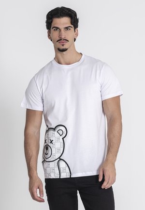 Camiseta de algodón blanca con un gráfico de oso de peluche contorneado en negro y patrones de logotipos repetidos en la parte inferior izquierda. Mangas cortas, cuello redondo.