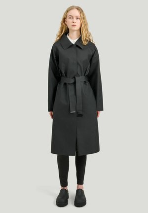 Nuori nainen, jolla on laineikas vaalea hiukset, pukeutuneena mustaan vyölliseen sadetakkiin, mustiin leggingseihin ja mustiin slip-on-kenkiin, seisoo yksiväristä taustaa vasten.