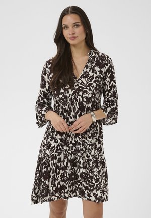 Žena s dlhými tmavými vlasmi, oblečená v čierno-bielych kvetinových šatách po kolená s tromi štvrťovými rukávmi a strieborným náramkom.